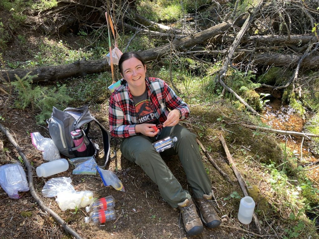 Samantha Motz sitting in the shade of a tree, labeling a water bottle for sampling.