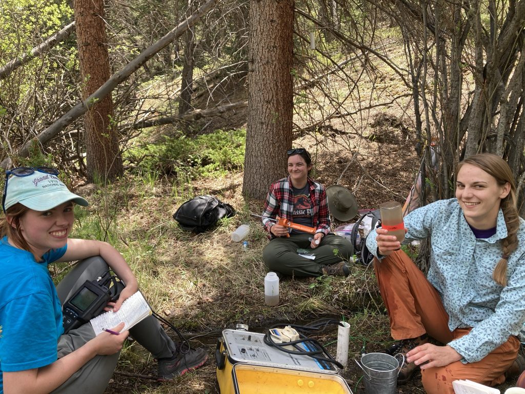 Sadie Jonson, Samantha Motz, and Sara Warix sitting in the shade under trees, smiling for the camera while they label bottles for water sampling.