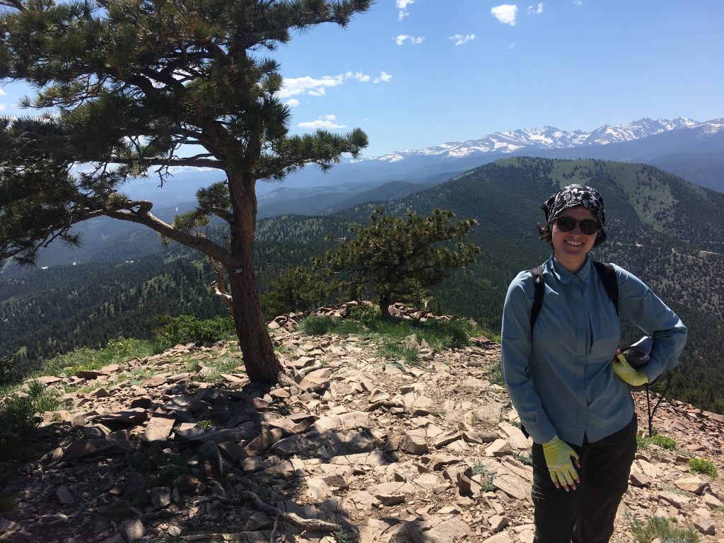 2021 RESESS Intern Shana Egan at the top of Sugarloaf Mountain in Colorado. There's a tree next to her and mountains visible in the background.