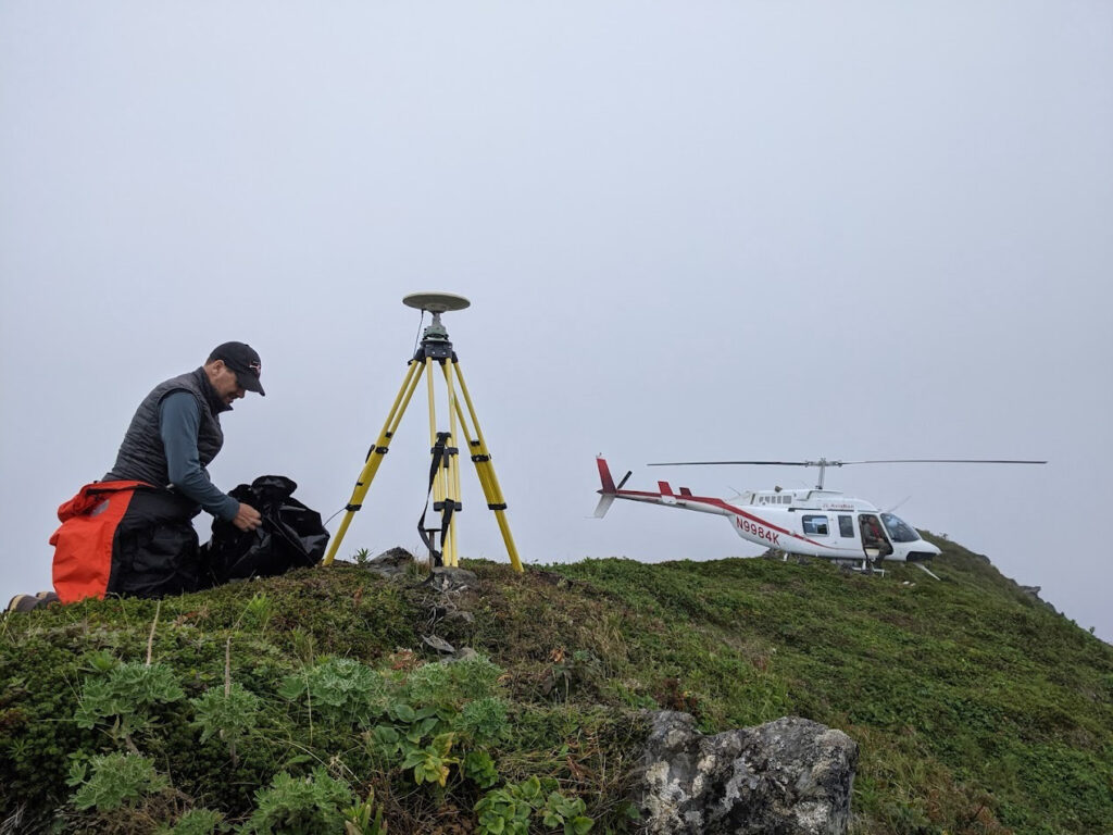 man kneeling beside instrument on tripod, helicopter on the ground behind