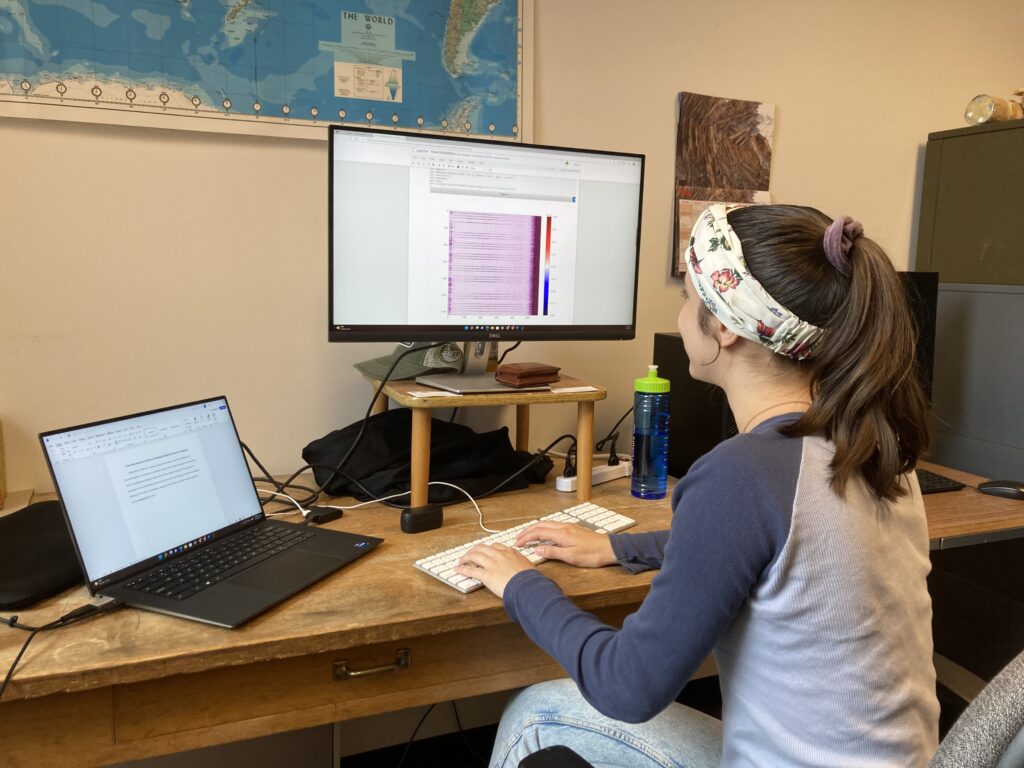2022 RESESS intern Halina Dingo types on a keyboard. The laptop to her left has a document open and the monitor to her right shows a purple-colored plot showing a vibroseis survey at a FORGE site.