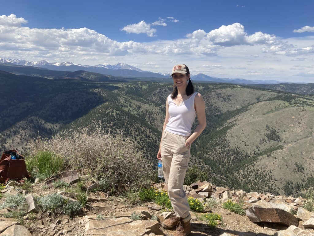 2022 RESESS intern Halina Dingo at the top of Sugarloaf Mountain in Boulder, CO. Rolling green hills are visible behind her with mountains on the horizon line.