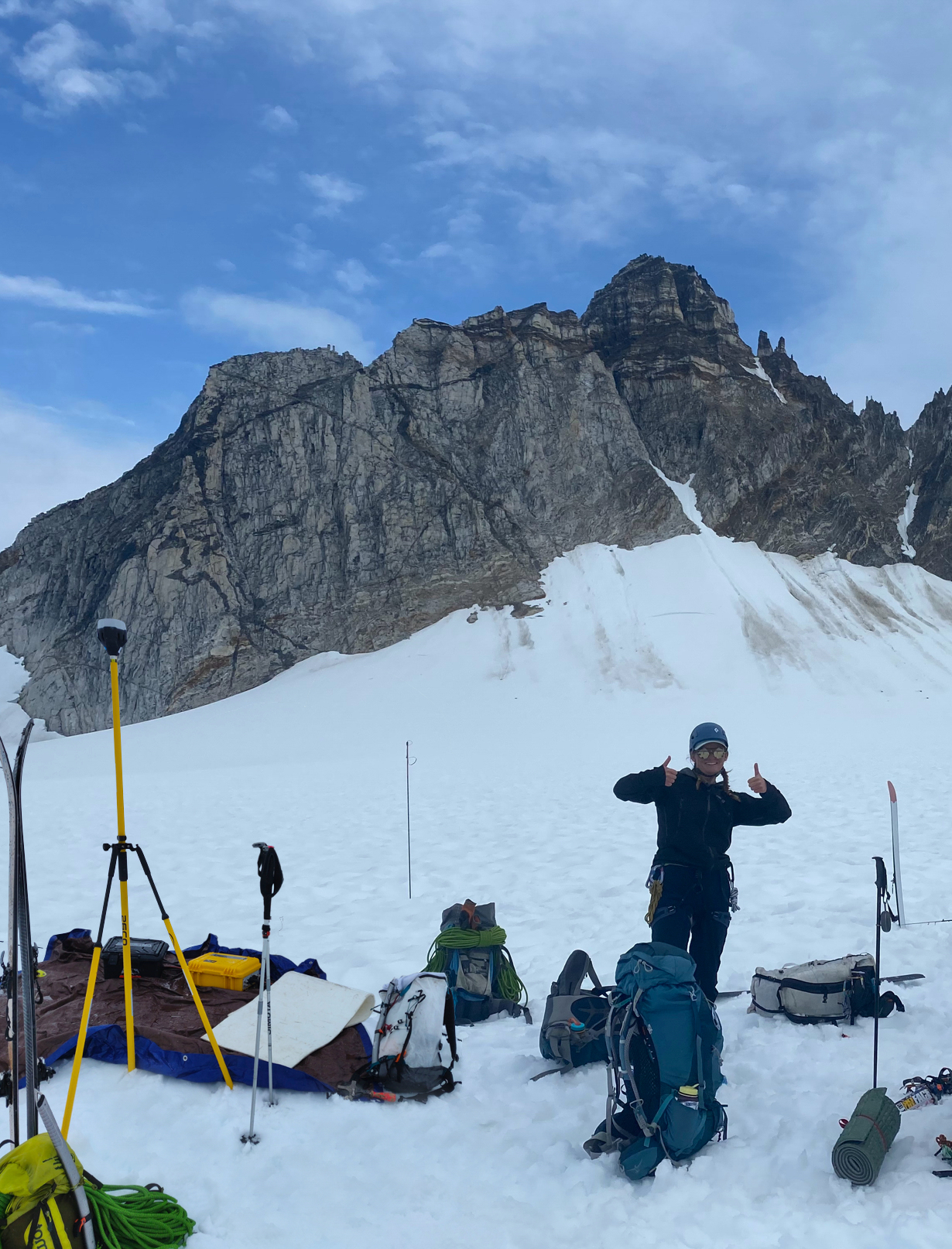 RESESS Satellite Intern Michela Savignano gives two thumbs up as she is surrounded by GPS equipment the Juneau Icefield Research Program in 2021. Snow covers the ground and there is a large outcrop behind her.