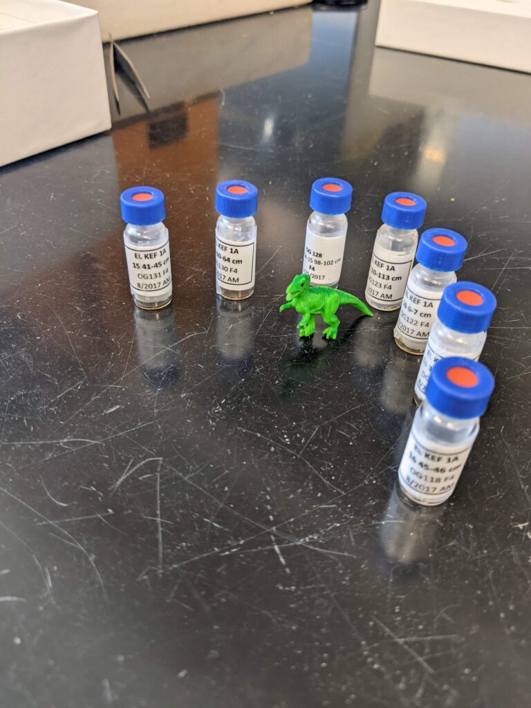 Seven samples in glass vials with blue and red caps surround a small, green T-rex figurine.