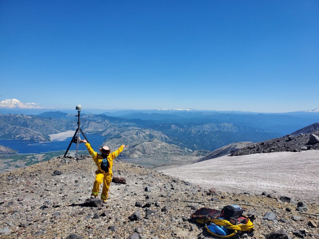 USIP Intern Kayla Byrd, in a bright yellow flight suit, poses in front of an upgraded GPS antenna and monument with Spirit Lake in the background on the Mount St. Helens campaign.