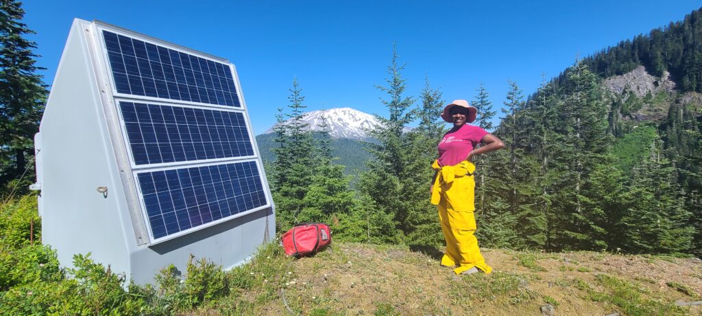 USIP Intern Kayla Byrd smiles next to solar panels with a snow-covered mountain in the background on the Mount St. Helens campaign trip.