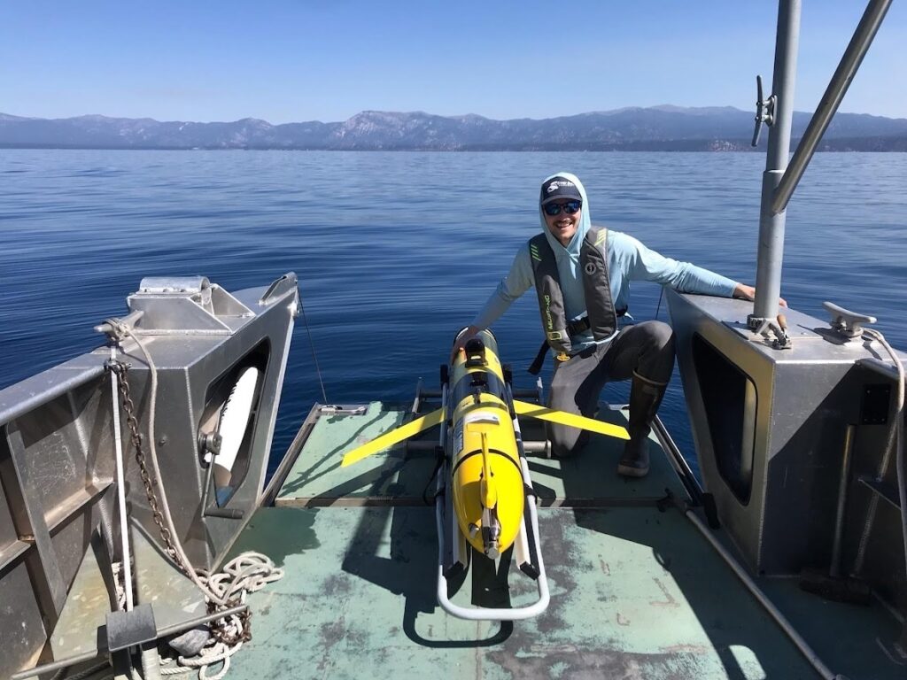 USIP Intern Kenny Larrieu is crouched next to an autonomous underwater vehicle on a boat, which he is about to deploy in Lake Tahoe.